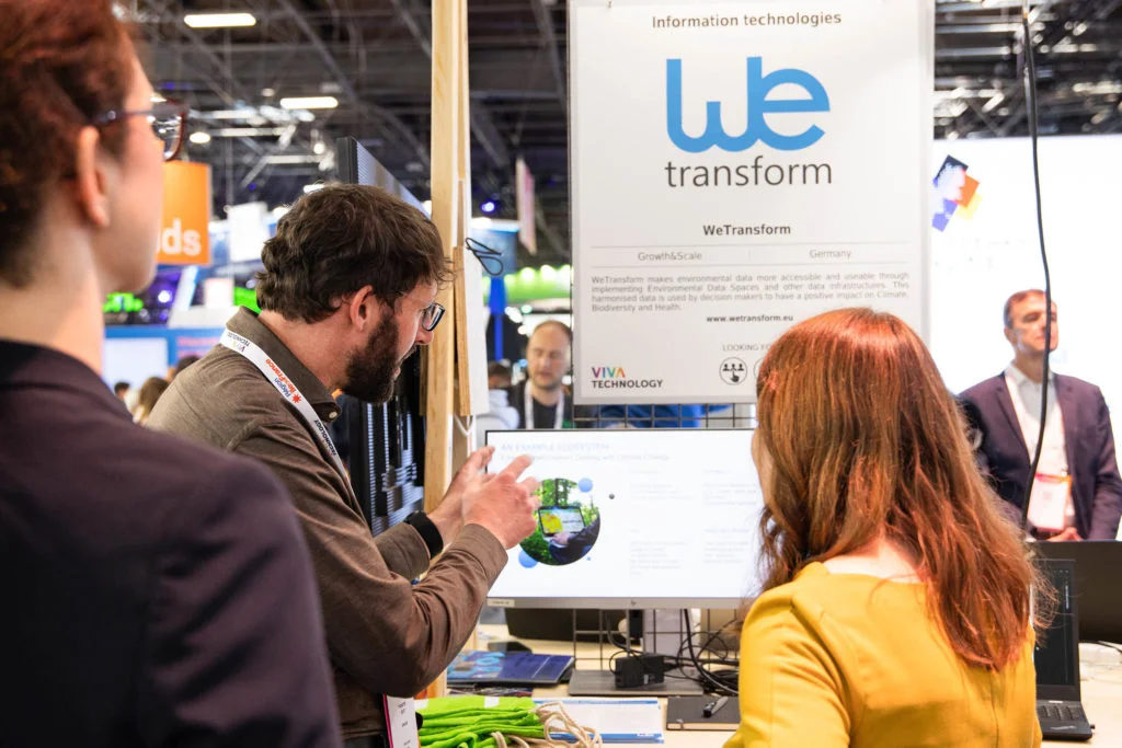 wetransform CEO Thorsten Reitz gestures at a screen underneath a wetransform poster at Viva Tech 2023 whilst explaining