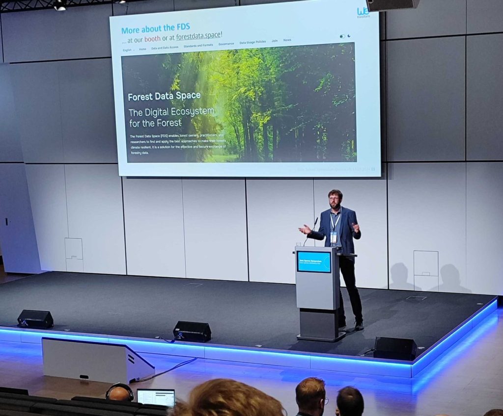 wetransform CEO Thorsten Reitz, a tall man sporting glasses, a mop of brown hair with a neat beard, and a casual blue suit. He is standing behind a digital lectern on a blue-lit stage, speaking to a crowd. Behind him is a large projection showing the homepage of the Forest Data Space - The Digital Ecosystem for the Forest.