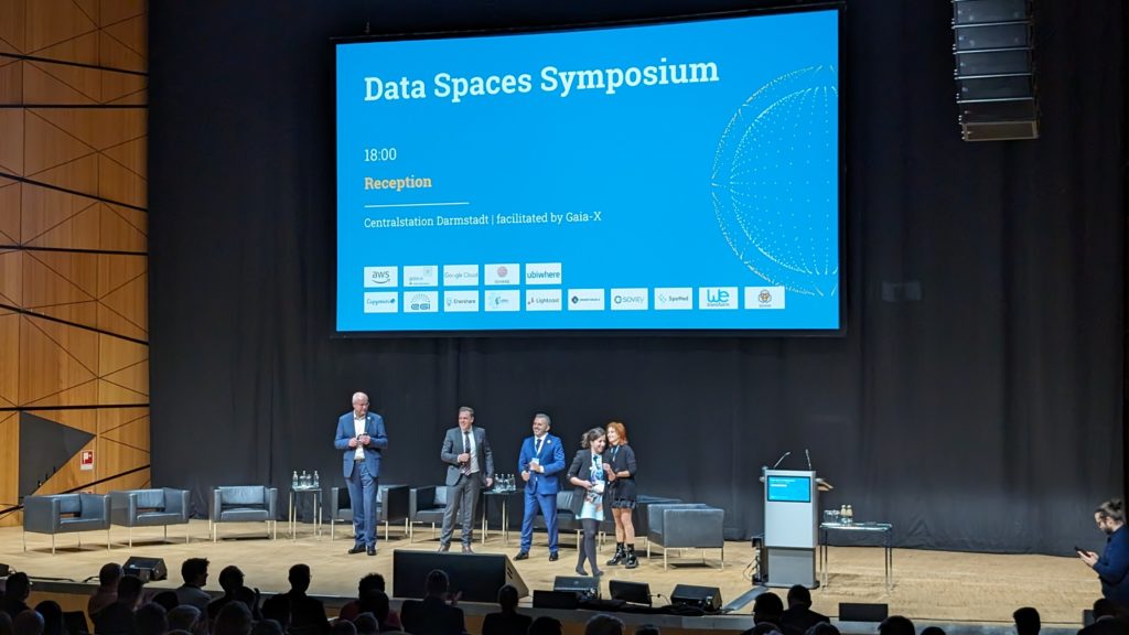 Five people are in the process of leaving a large stage, smiles on their faces. Behind them is a projected blue slide that reads "Data Spaces Symposium", along with the time and place for the reception and sponsor-logos.