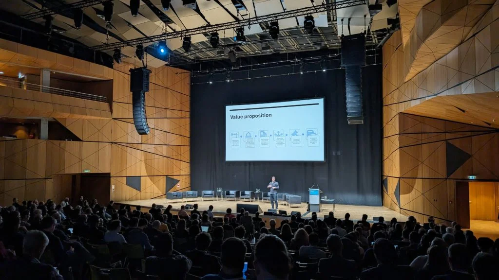 A dark-haired man in a black button-down shirt and jeans looks out at a seated crowd in a darkened conference theatre. Behind him are nine leather chairs, three tables with water, a lectern, and a large projection screen. On the screen is a slide titled "Value proposition"