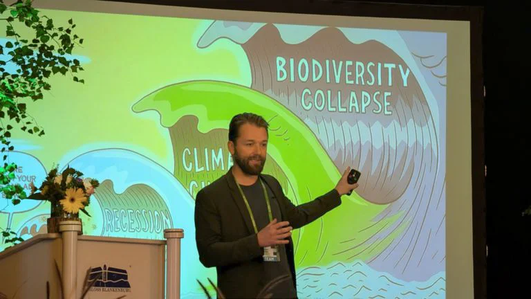 A bearded presenter in a dark outfit stands in front of a slide displaying three waves labelled biodiversity collapse, climate change, and recession