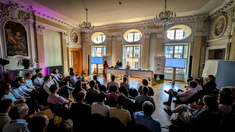 A seated crowd faces two presenters in a beautifully decorated German room at the Future Forest Forum 2024. Two chandeliers hang from the ceiling and centuries-old paintings are elaborately framed in light-coloured moulding. In front of the presenters is a long table and they are flanked by two monitors.