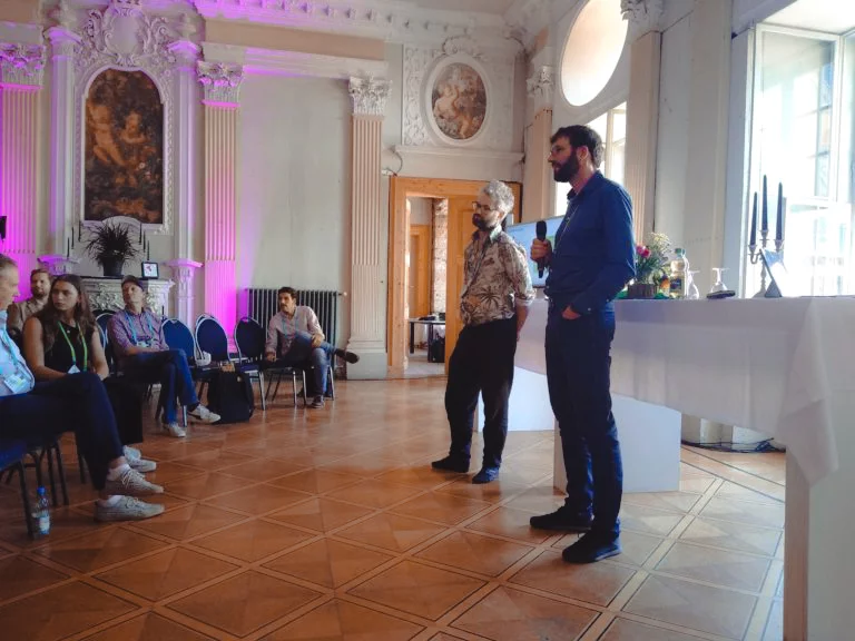 Two men stand on the right of the image, one holding a microphone. To the left, a seated crowd engages in lively discussion.