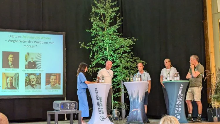 Five people stand on a stage. In front of them are three tables bearing the text "Future Forest Initiative". Behind them is a small tree. A projector in the foreground projects onto a screen to their left, featuring pictures of all five people under the header "Digitaler Zwilling des Waldes - Wegbereiter des Waldbaus von morgen?"