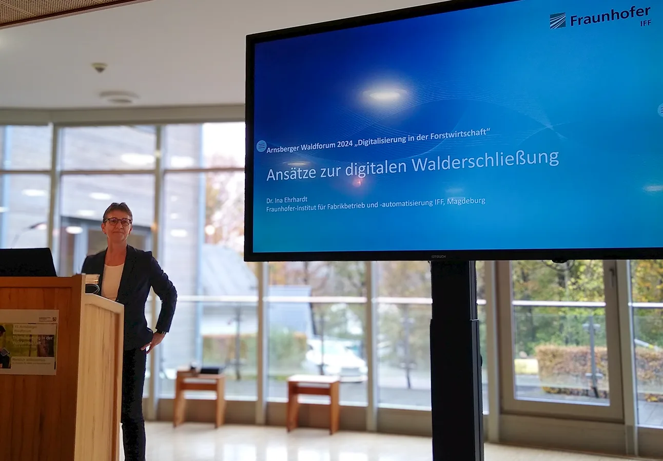 Dr. Ina Ehrhardt of Fraunhofer IFF stands behind a wooden lectern at Arnsberger Waldforum 2024. There is a glass wall behind her. Beside her is a large monitor showing the blue opening slide of her presentation "Ansätze zur digitalen Walderschließung"
