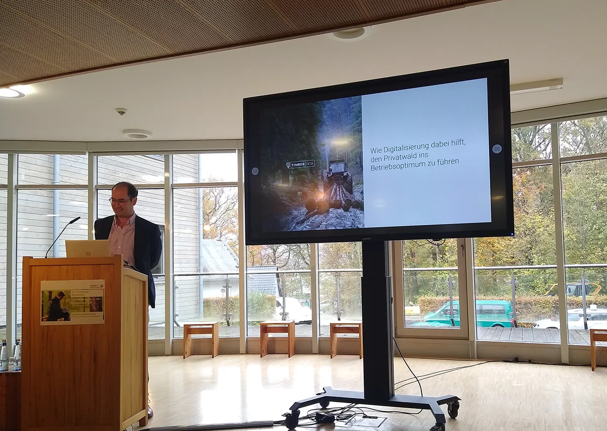 Ludwig Graf Douglas of TimberTom presents from behind a wooden lectern. Behind him is a wall made of windows, showing a partial view of a wood-clad building and a small parking lot among many trees and shrubberies. Next to him is a large monitor showing a picture of forest machinery with the text "TIMBERDATA" on the left and a white square with the green text "Wie Digitalisierung dabei hilft, den Privatwald ins Betriebsoptimum zu führen" on the right.