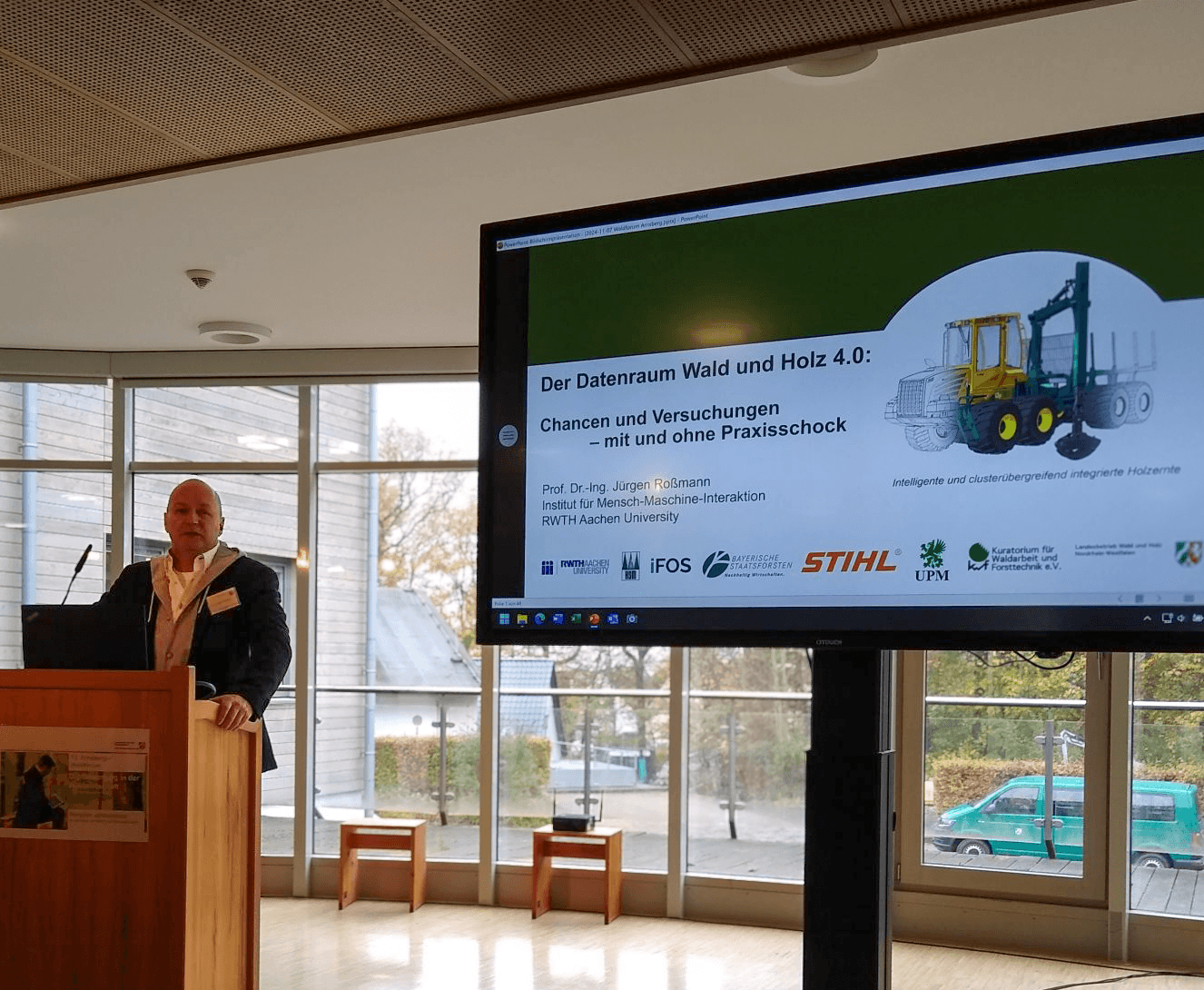 Prof. Dr. Jürgen Rossmann of RWTH Aachen stands at a wooden lectern bearing Arnsberger Waldforum branding. To the right is a large monitor showing the opening slide of his presentation "Der Datenraum Wald und Holz 4.0: Chancen und Versuchungen - mit und ohne Praxisschock"