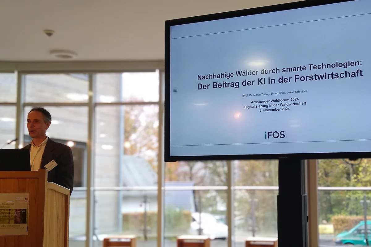 Prof. Dr. Martin Ziesak of iFOS presents from behind a wooden lectern. A large monitor beside him shows the opening slide of his presentation "Nachhaltige Wälder durch smarte Technologien: Der Beitrag der KI in der Forstwirtschaft"