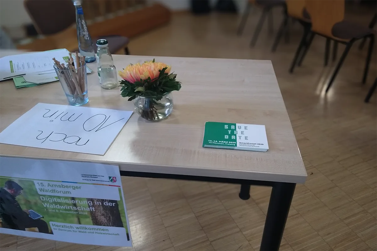 A stack of green/white SmartForest 2025 "Save the Date"-flyers sits on a table from which hangs a welcome sign to the 15. Arnsberger Waldforum. Also on the table are some flowers, two empty glass bottles, a glass filled with wooden ballpoint pens, a stack of conference programmes, and a handwritten sign reading "noch 10 min" in loopy handwriting.