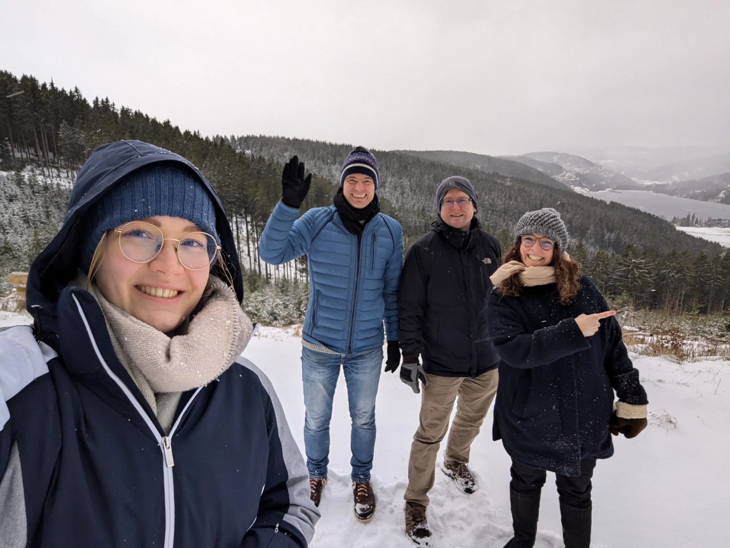 A selfie taken by team members on a snowy walk, with everyone bundled up tightly. Julia, a young blonde woman, takes the selfie. To her right is Chris, waving at the camera. Next to him, a smiling Simon. Finally, Johanna grins broadly as she points at the gorgeous lake nestled in the woods behind them.