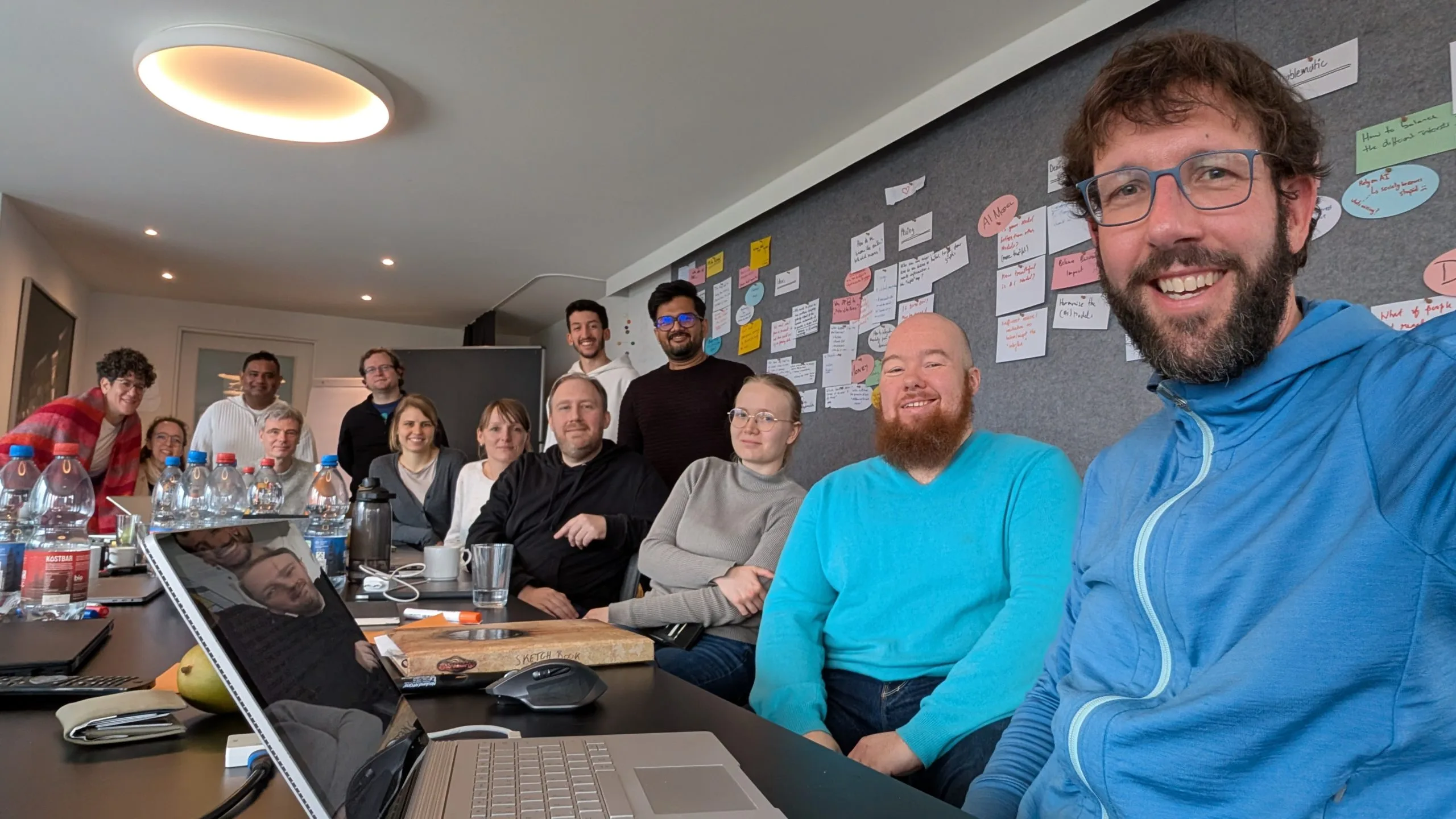 The wetransform team takes a selfie in the meeting room. Flaminia, Johanna, Somakanthan, Christopher, Simon, Anna, Franziska, Morchid, Florian, Kapil, Julia, John, and Thorsten all smile at the camera whilst gathered around the conference table. In the background, a felt wall is full of notes.