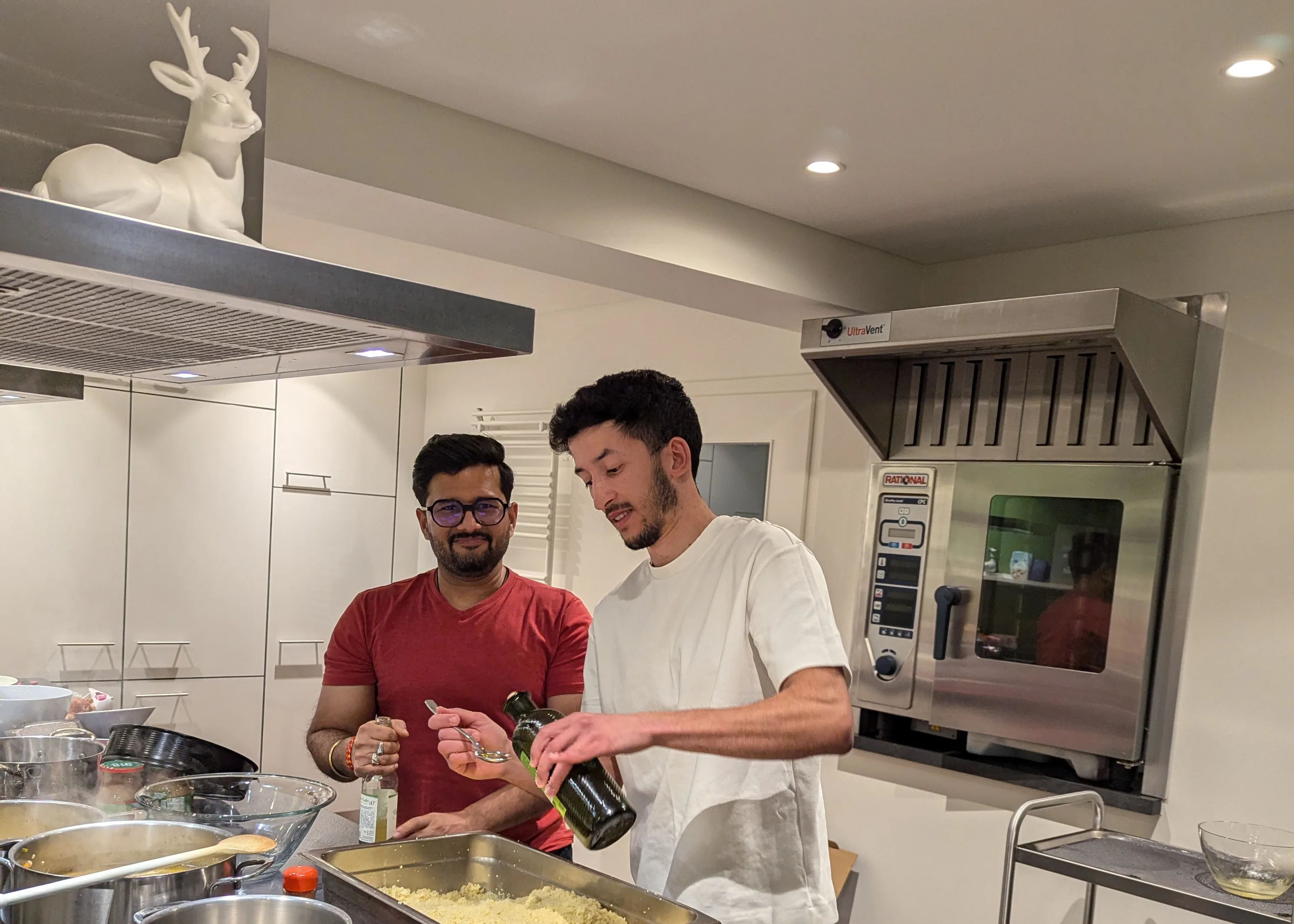 Kapil and Morchid of the wetransform team cook together in a large commercial kitchen. Kapil, a bearded man in glasses, smiles at the camera as Morchid, a bearded man without glasses, pours liquid from a green bottle onto a spoon.