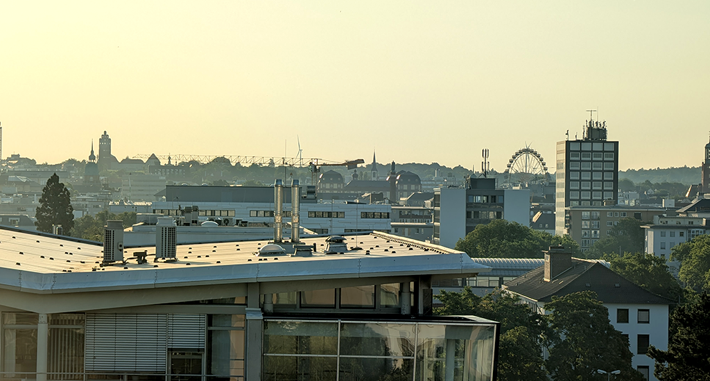 A view of across the roofs of Darmstadt, taken from the northwest of town, facing southeast towards the centre. Green trees, buildings of different ages and styles, construction cranes, windmills, the distinctive hand-shape of the Hochzeitsturm at UNESCO World Heritage Mathildenhöhe, and a Ferris Wheel can be seen against a summer sky.
