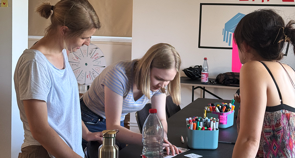 Three women from the wetransform team are gathered around a black table on which there are two blue bins full of markers. Anna, a blonde sporting a white shirt, looks on from the left. Julia, also blonde, wearing a striped top and jeans, leans over as she writes something on a card. To the right, Ramona's dark hair and colourful dress are seen from the back.