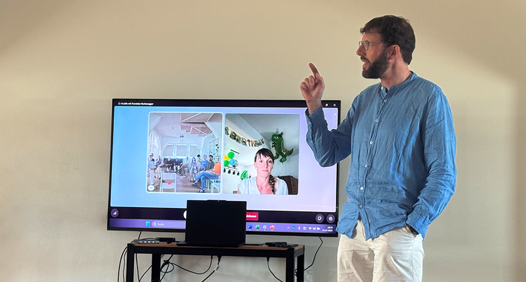 wetransform CEO Thorsten Reitz stands next to a large monitor. He has dark hair, glasses, and a beard, and is sporting a blue button-up shirt and light trousers. His finger is raised and his mouth slightly open as he speaks. On the monitor is a videocall, showing the bulk of the wetransform team gathered in the offsite space on the left and Lead Project Manager Franziska to the right, with a birthday banner and green dinosaur balloon in the background.