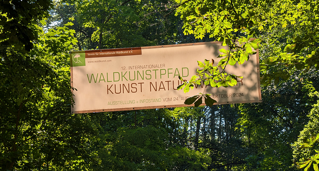 A large banner hangs high between sun-dappled green tree foliage. It reads "12. Internationaler Waldkunstpfad. Kunst Natur Wasser".