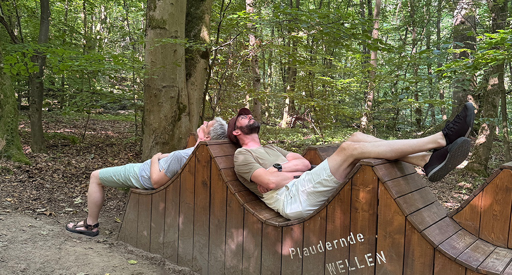 Two men from the wetransform team lay down back-to-back across a wave-shaped wooden construction in the woods. Lead Sales Chris stretches out to the left, his grey hair just touching CEO Thorsten's brown hat. Thorsten lays in the opposite direction, his legs up over the crest of the "wave". On the structure is the white text "Plaudernde WELLEN".