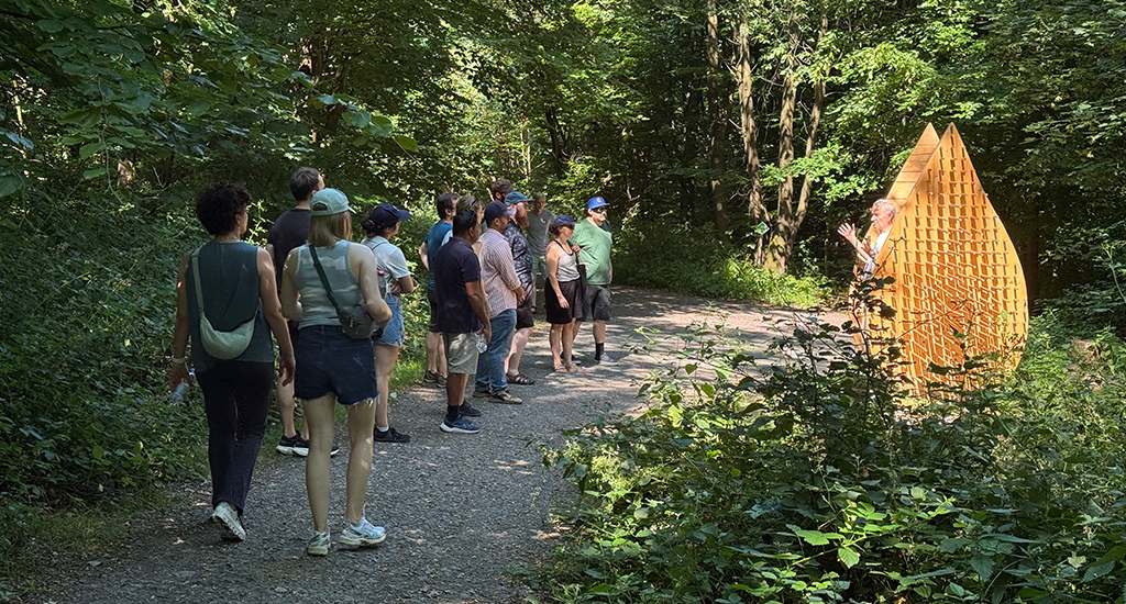 The wetransform team is on a forest path. They are looking intently at the Waldkunstpfad tour guide, who stands in a sunny spot between two wooden constructions shaped like water drops, his hands spread as he explains the artist's vision.