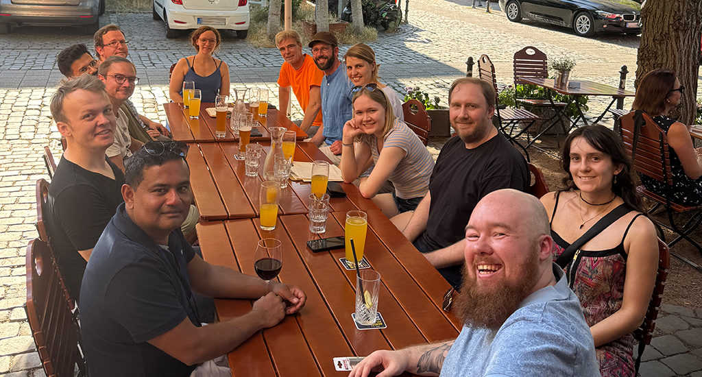 A restaurant's terrace on a cobbled street in Darmstadt on a sunny day. Thirteen members of the wetransform team sit at three combined wooden tables with a variety of drinks on it. Everyone is smiling at the camera.