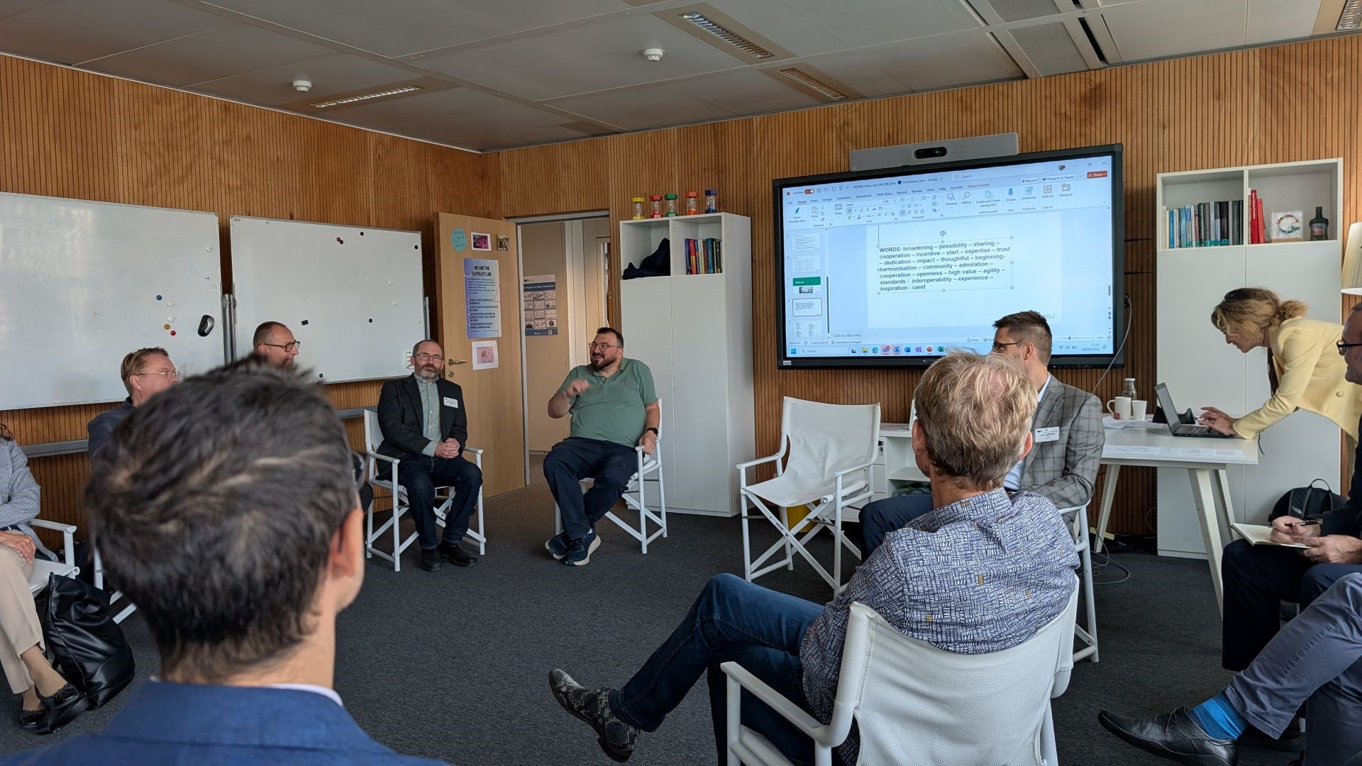 A group of eleven people in a wood-panelled room. Ten are seated on grey folding chairs, while one stands to the right of the picture, typing on a laptop. Central is a large monitor, showing a PowerPoint presentation with words people associate with INSPIRE.

