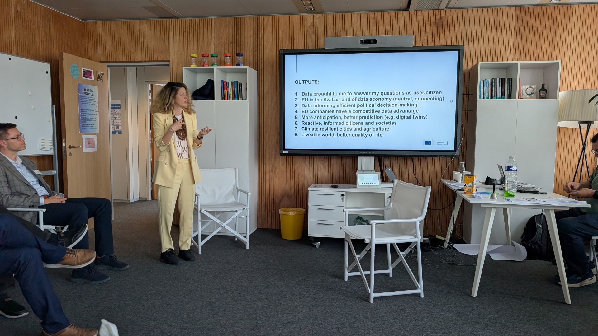 A blonde woman in a yellow suit stands next to a large screen, presenting. In the bottom right of the screen is the logo of the European Commission. The slide's title reads "OUTPUTS:", which is followed by a list of eight items.