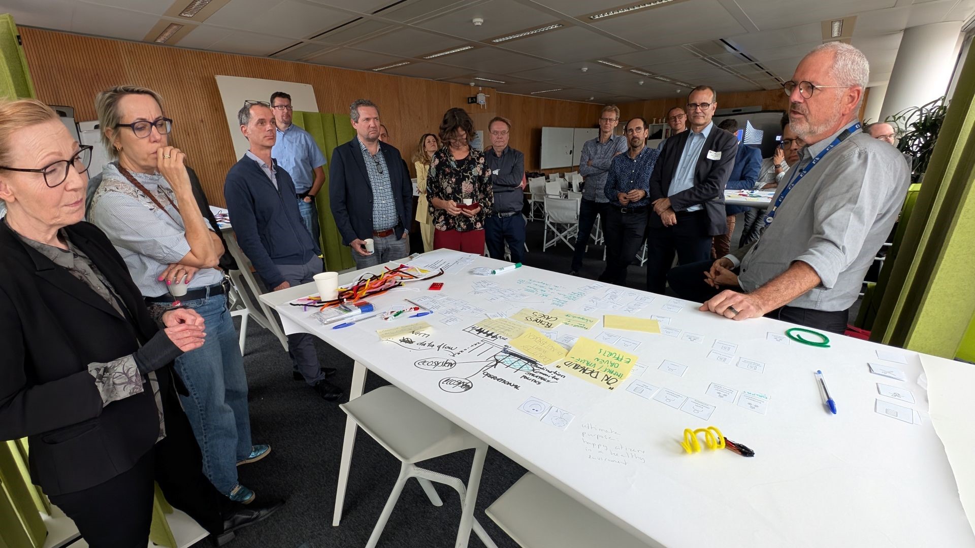 A group of 18 people is gathered around a white table, where a man in glasses and a grey button-down shirt is explaining something. On the table are many different bits of paper, several pens, and a handful of colourful pipe cleaners. There is also writing and a drawing directly on the table's surface. Words such as "ecosystem", "data flow", "strategy/politics", and "operational" stand out.