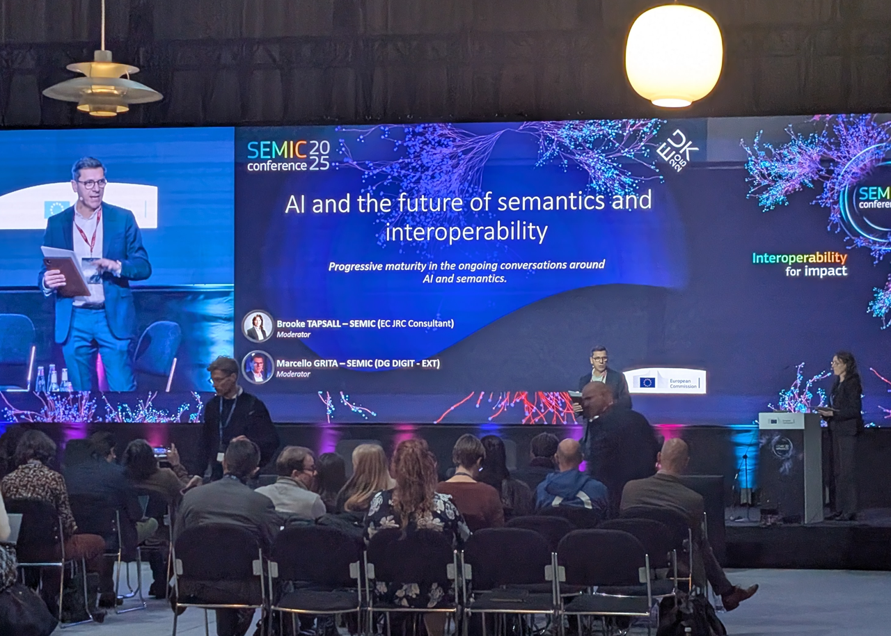 Photograph taken at the SEMIC 2025 panel "AI and the future of semantics and interoperability - Progressive maturity in the ongoing conversations around AI and semantics.". A group of people is seen from the back, seated on rows of black chairs facing a stage. On the stage are two people, identified on the screen behind them as the session's moderators Brooke Tapsall (EC JRC Consultant) and Marcello Grita (DG DIGIT - EXT)