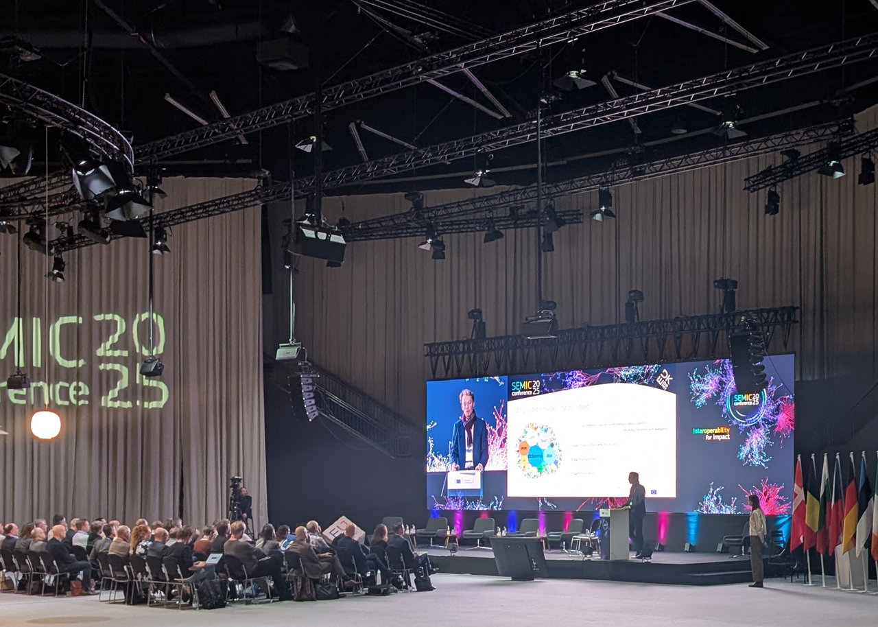 Photo taking from the back of the room at SEMIC 2025. Black metal beams run across the equally black ceiling, supporting many lamps and all sorts of tech. The walls are draped in a grey fabric, onto which the words "SEMIC Conference 2025" is projected. A large group of people sit in dark chairs, watching the stage, where someone is presenting. Their slides as well as a close-up view of them are visible on a large screen behind them. To the right of the stage stand the flags of the EU nations.