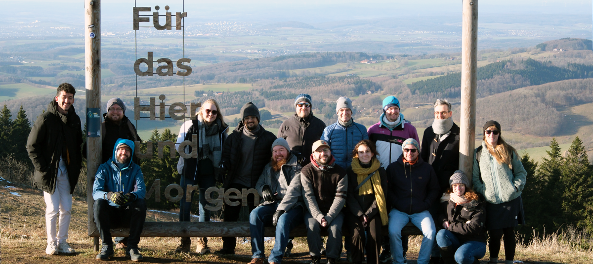 Group photo from the 2026 wetransform offsite. Fifteen people pose inside a wooden frame, with the German text 