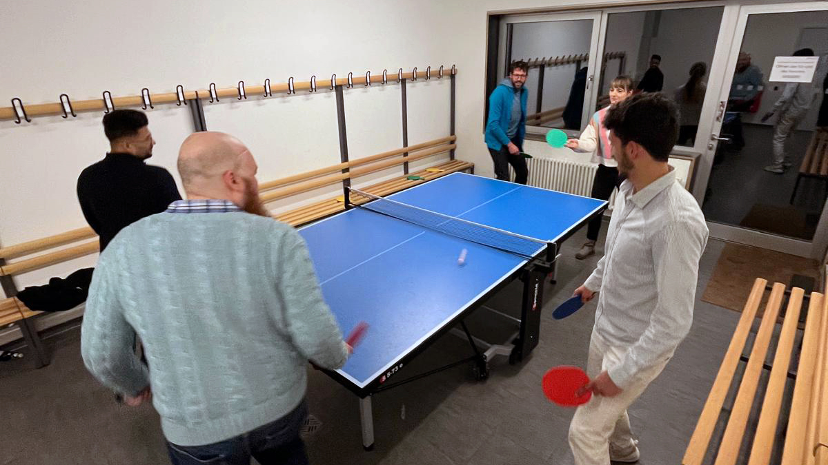 Five members of the wetransform team, Kapil, Thorsten, Franziska, Morchid, and John, are dashing around a blue table tennis table, bats in hand. The white ball is mid-bounce in the centre.