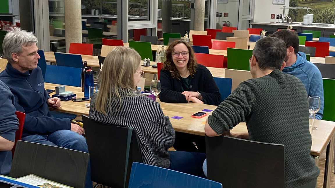 Five members of the wetransform team: Chris, Julia, Johanna, Thorsten, and Nathanael are gathered at a wooden table with colourful chairs in a canteen, laughing as they engage in a game with small purple cards.