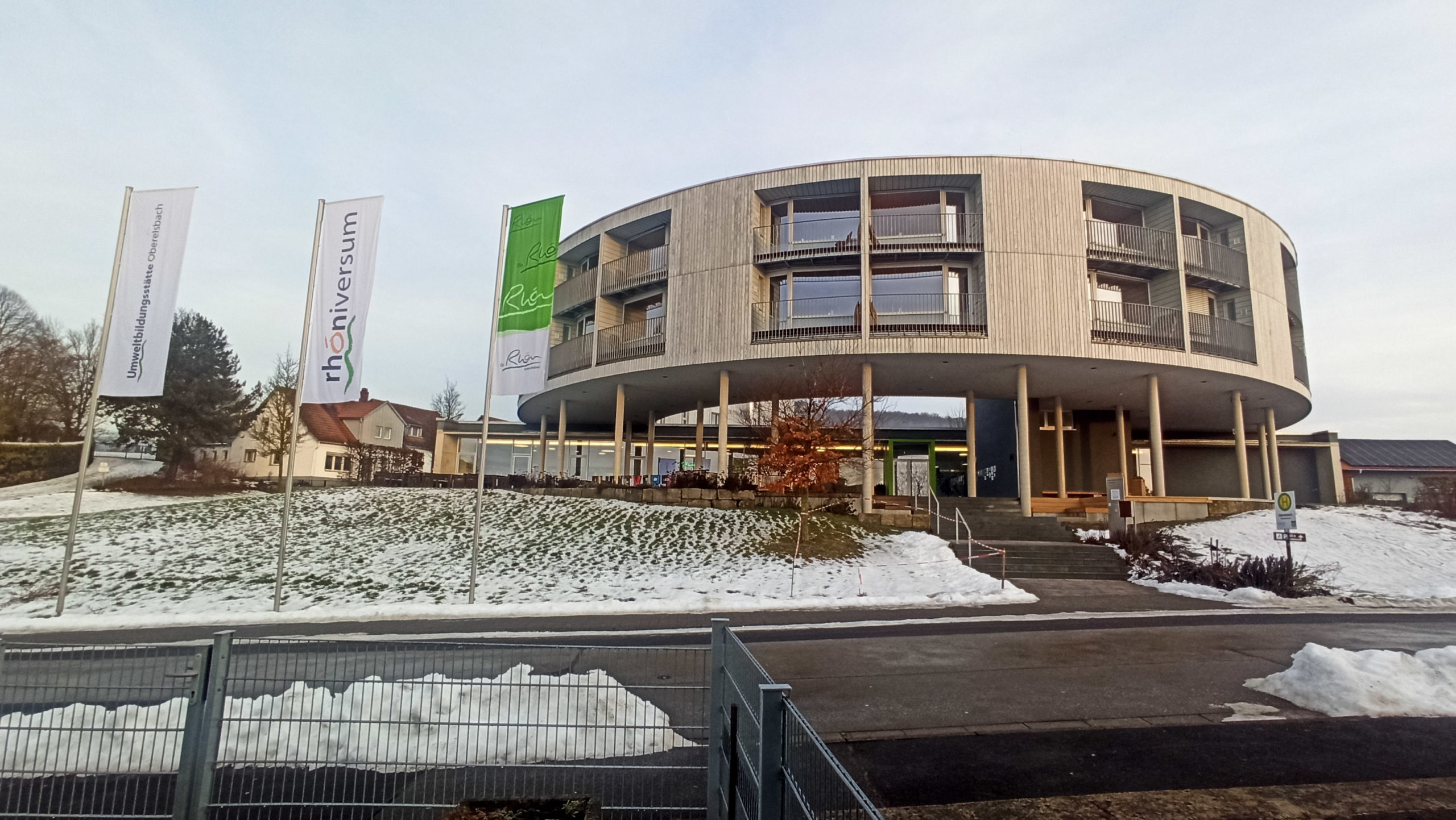 The Rhöniversum, a large modern greyish building with a long rectangular ground floor. In front is a half-circle of pillars that partially holds up two circular-shaped floors above.There are many large windows and balconies recessed into the building. The ground is covered in snow and to the left are three flagpoles, displaying flags that read: "Umweltbilldungsstätte Oberelsbach", Rhöniversum", and "Rhön".