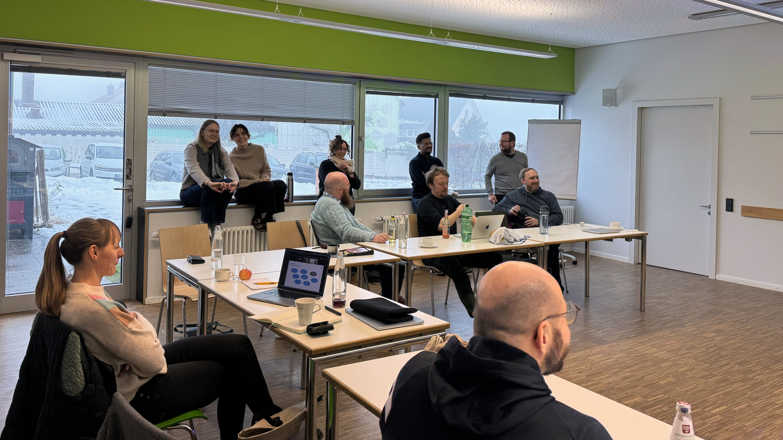 Part of the wetransform team inside a classroom at the Rhöniversum during the 2026 offsite. Half the room is visible, with a wooden floor and walls in white and green. There are large windows as well as a glass door, showing snow outside. Five desks are loosely arranged in an L-shape and ten people are talking and smiling. Some sitting in chairs, some perched on the windowsills by the radiators.