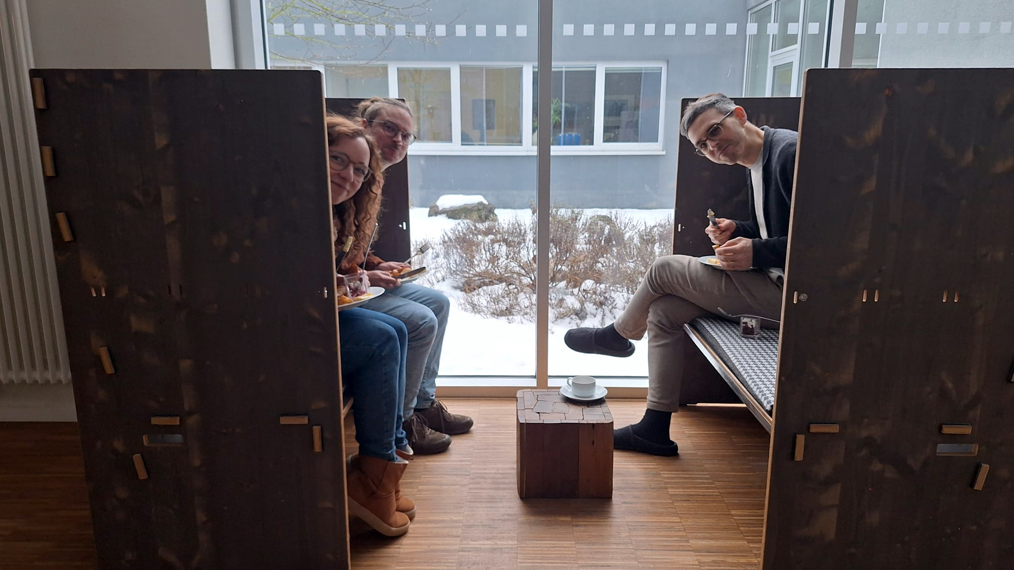 wetransform team members Johanna, Fabian, and Nathanael peek out from the wooden booth in which they are having lunch, plates perched on their laps and cutlery in their hands, and smile at the camera. In the background is a large window, showing a small patch of snow-covered plants and rocks and the outside of a grey building with many windows.