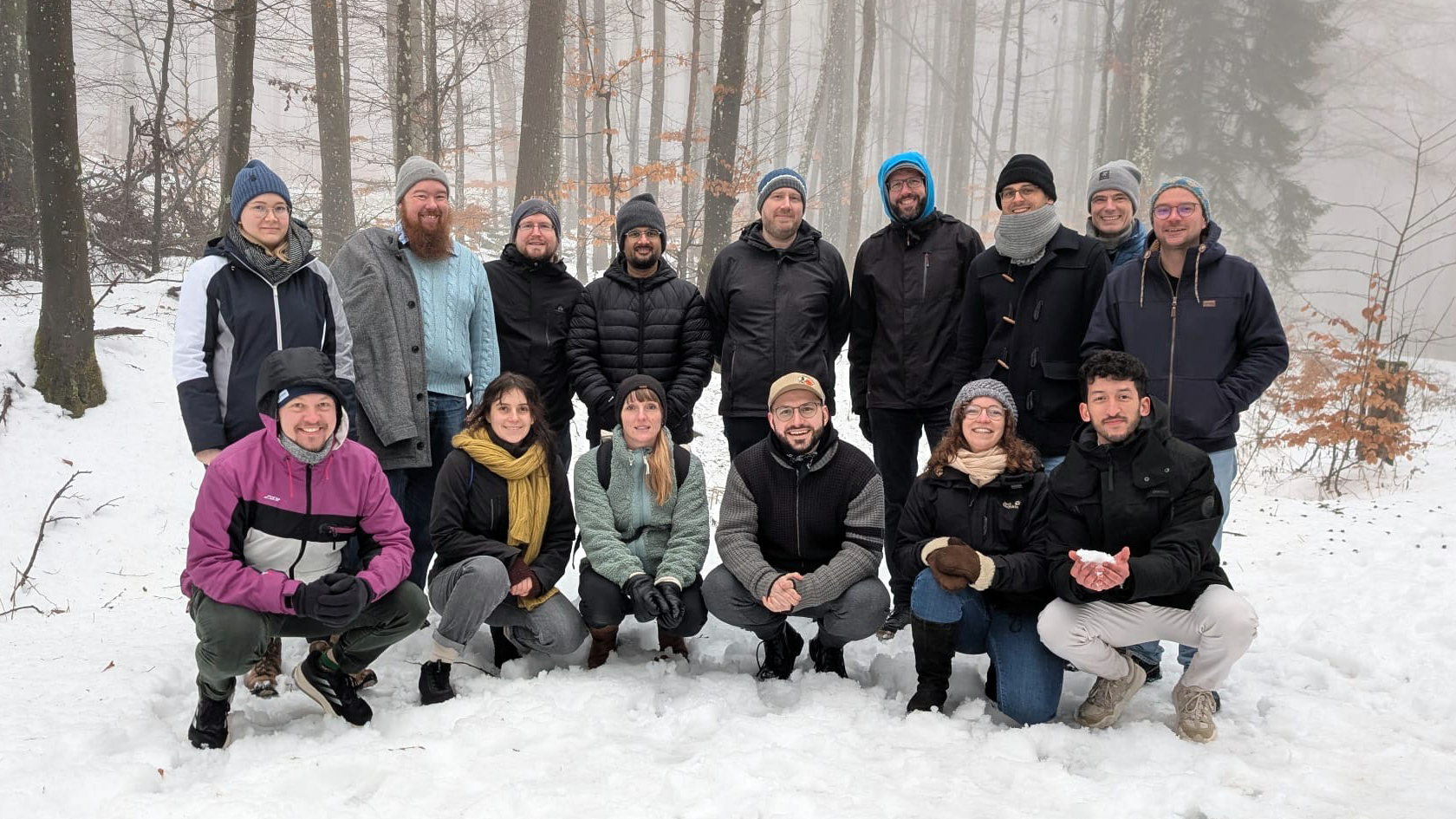 Group photo from the wetransform 2026 offsite. Fifteen people pose in a snowy forest, nine standing in the back, six squatting in front. All smile at the camera.