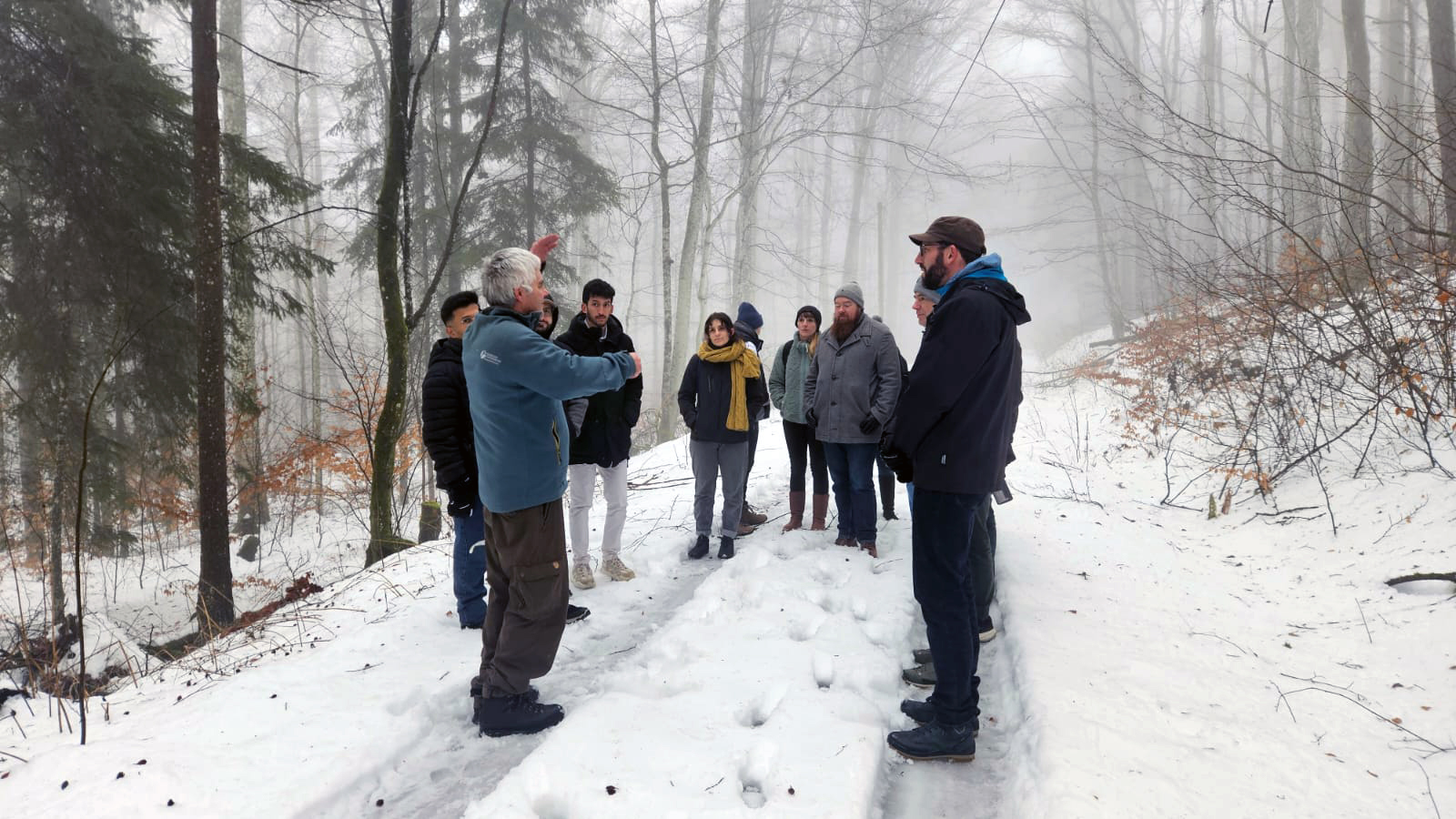 A grey-haired forester in a blue sweatshirt stands amid the wetransform team in a snowy forest, gesturing as he explains something to the crowd.