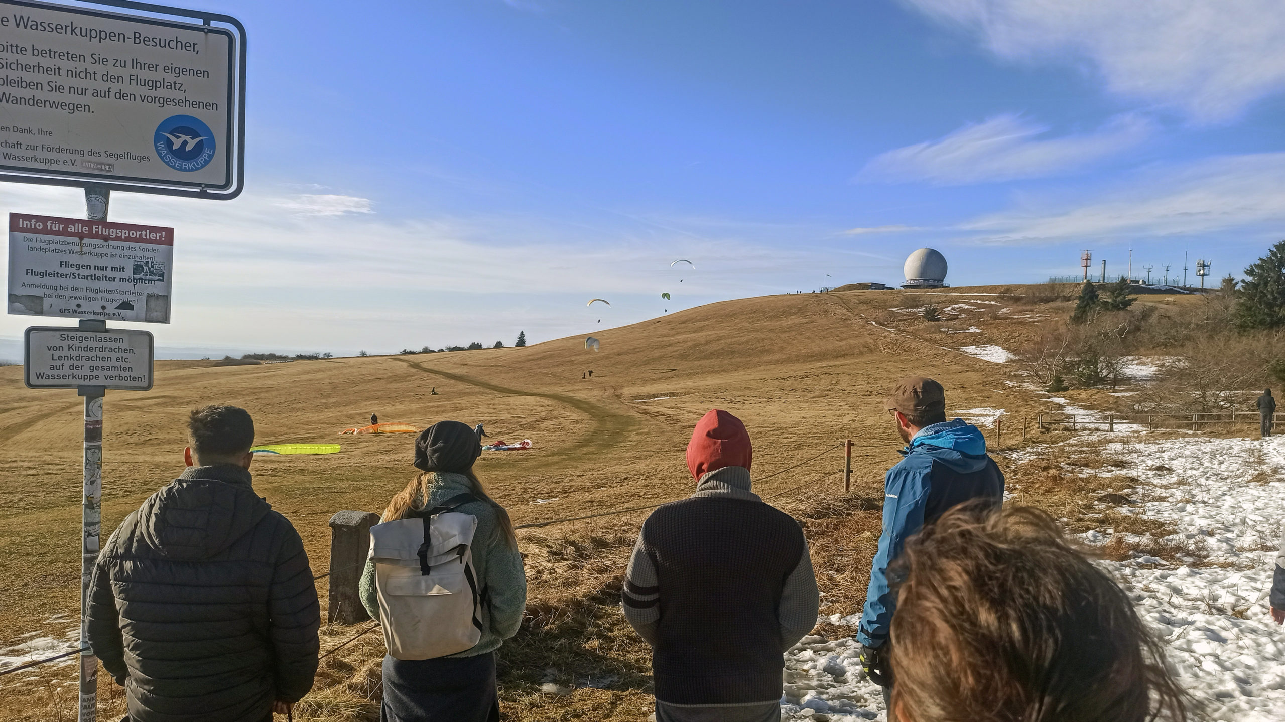 Five members of the wetransform team are seen from the back as they look towards a large hill with an old radar dome on it. On the field between the group and building are people engaging in air sports. Four actively paragliding and three still on the ground. To the left is a signpost with three elaborate German signs about air sport safety, one of which is sporting the blue Wasserkuppe logo.