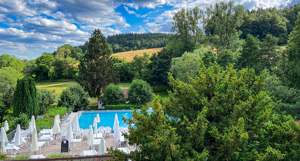 View of a pool with a terrace, surrounded by wooded hills and large swathes of grass under a blue sky with some clouds.