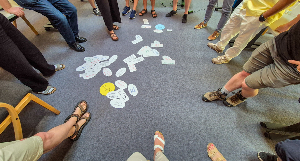 A downward shot of a grey floor. A large amount of handwritten notes are central, combined in small clusters. Surrounding these, the legs and feet of thirteen people are visible.