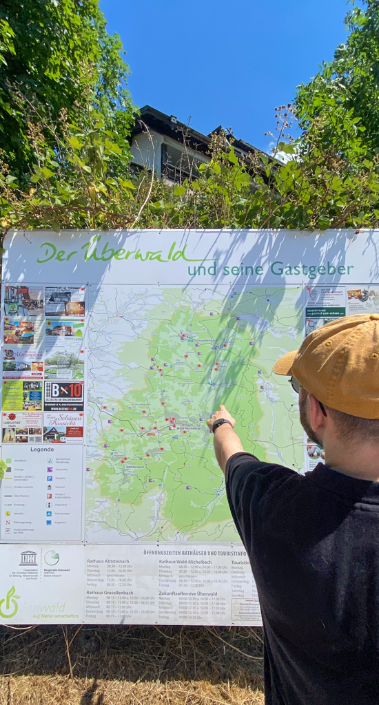 A bearded man in a yellow hat, black shirt, and glasses is seen from behind as he points at a large map outside. Above the map, it reads "Der Überwald und seine Gastgeber"