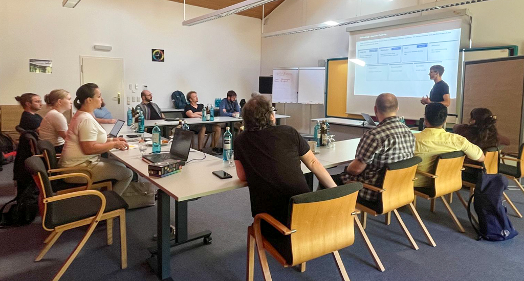 The wetransform team is seated around a U-shaped combination of tables in a bright meeting room, all looking at a projection screen in front of which Nathanael is presenting.
