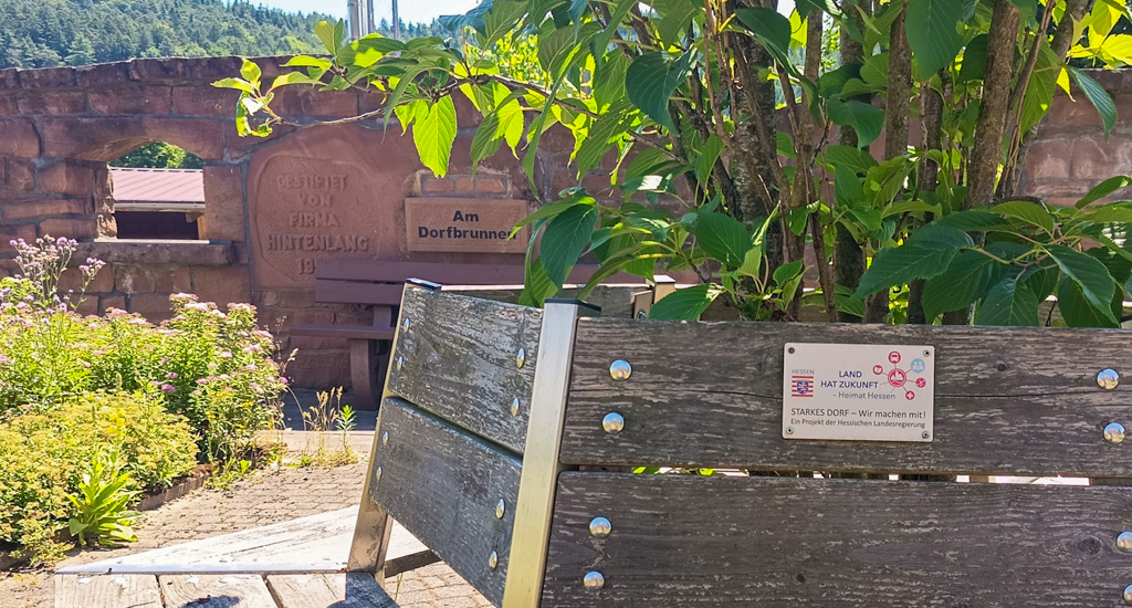 Benches surround a young tree on a sunny day. On the foremost bench is a new metal plaque bearing the Hessian coat of arms and the text "LAND HAT ZUKUNFT - Heimat Hessen", followed by an image showing stylised connections between people, nature, knowledge, and transport, and finally the text "STARKES DORF - Wir machen mit! Ein Projekt der Hessischen Landesregierung". In the background is another bench, with a sign above it that reads "Am Dorfbrunnen" in clear black lettering. Embedded in the same old wall the sign is on, there's another text raised into the stone "GESTIFTET VON FIRMA HINTENLANG 19", with the final numbers disappearing behind the bench.