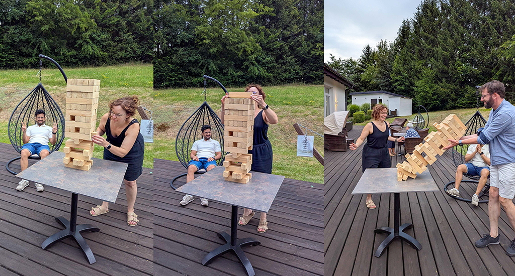 A combination of three photographs: 1 - Johanna tests blocks in a Jenga tower almost as tall as she is while Kapil flashes peace signs at the camera in the background. 2 - Johanna places a block atop the tower. 3 - Thorsten attempts to catch the tower as it falls, while Johanna and Kapil look on, laughing