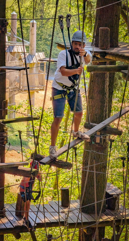 Kapil, an Indian man with a beard and glasses, is all rigged up in safety gear as he climbs along a narrow plank suspended by ropes in between trees during an outing at a Kletterwald ("climbing forest").