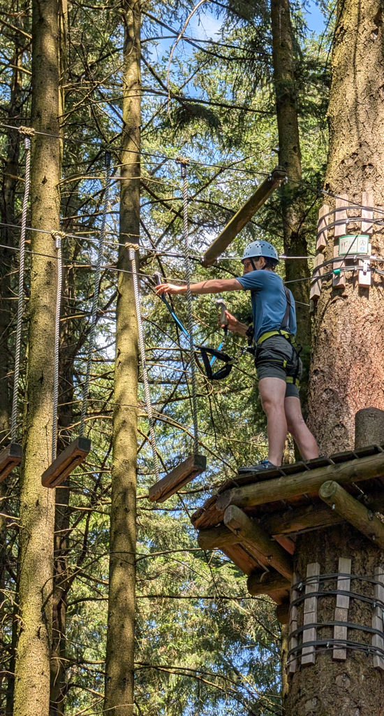 Nathanael, a tall white man in glasses, is decked out in a safety harness and helmet as he secures himself to a cable between trees during an outing at a Kletterwald ("climbing forest")